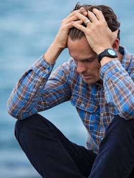 A man in a plaid shirt sits by the water looking distressed, symbolizing stress.